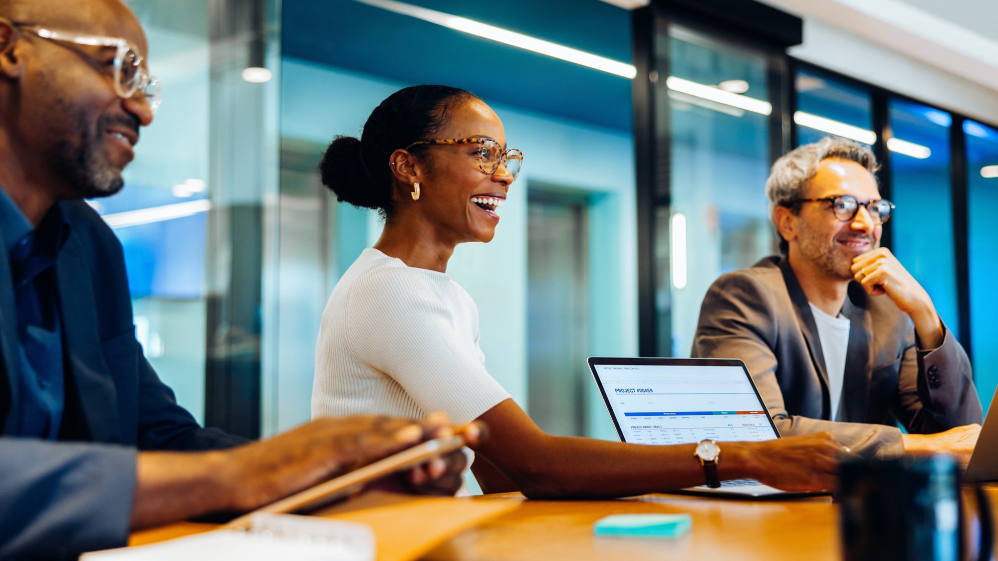 Three diverse coworkers, smiling and engaged in discussion around a meeting table in an office setting, collaborating with laptops and notes.
