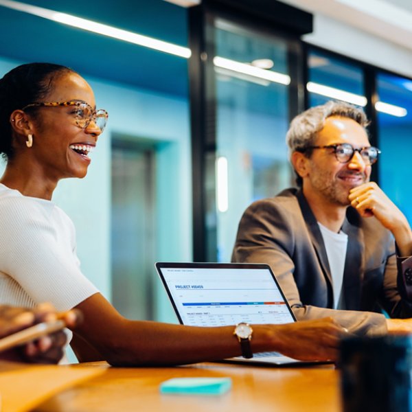 Three diverse coworkers, smiling and engaged in discussion around a meeting table in an office setting, collaborating with laptops and notes.