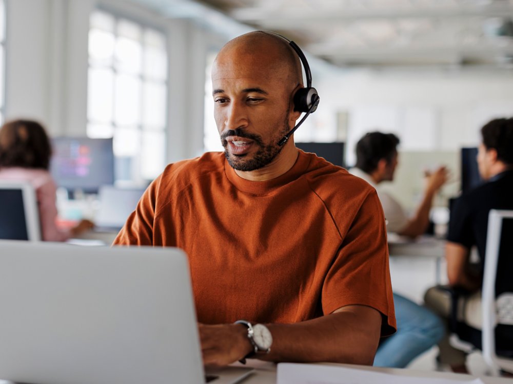 Customer service agent is handling a call while typing on his laptop in a busy office