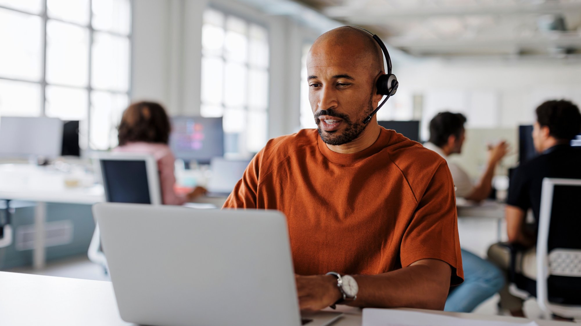 Customer service agent is handling a call while typing on his laptop in a busy office