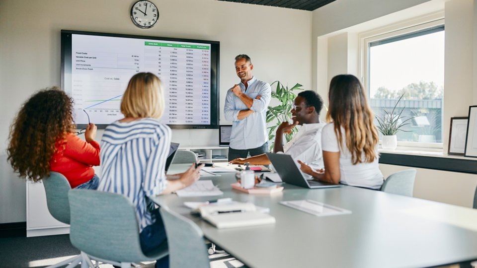 A diverse group of colleagues engaged in a lively discussion during a presentation in a modern office setting. The teamwork and professional atmosphere are emphasized through charts and collaborative interaction.