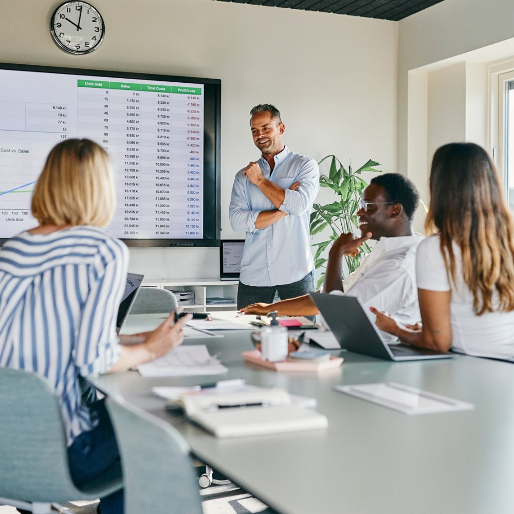 A diverse group of colleagues engaged in a lively discussion during a presentation in a modern office setting. The teamwork and professional atmosphere are emphasized through charts and collaborative interaction.