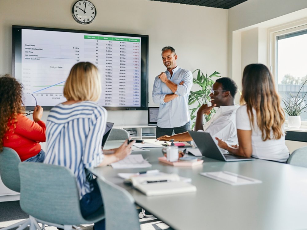 A diverse group of colleagues engaged in a lively discussion during a presentation in a modern office setting. The teamwork and professional atmosphere are emphasized through charts and collaborative interaction.