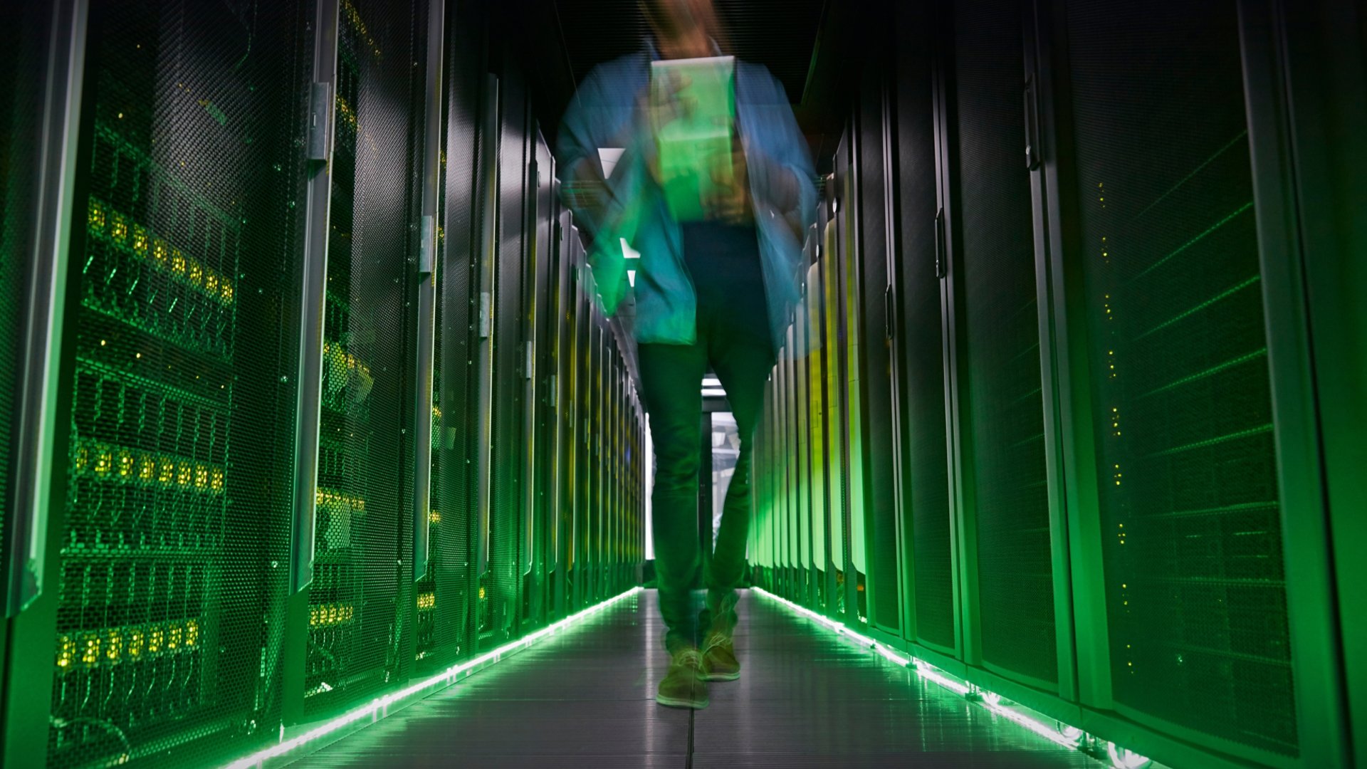 A person walks down a narrow aisle between tall server racks in a data center. The motion blur suggests fast movement or busy activity in the high-tech environment. Green lighting and indicator LEDs create a futuristic glow along the floor and equipment. The scene emphasizes modern infrastructure, digital networks, and information technology operations.