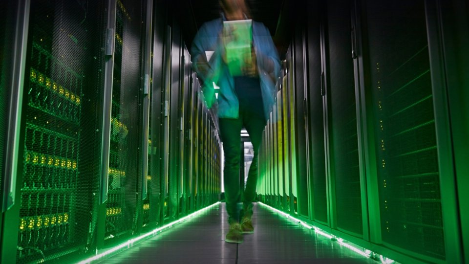A person walks down a narrow aisle between tall server racks in a data center. The motion blur suggests fast movement or busy activity in the high-tech environment. Green lighting and indicator LEDs create a futuristic glow along the floor and equipment. The scene emphasizes modern infrastructure, digital networks, and information technology operations.