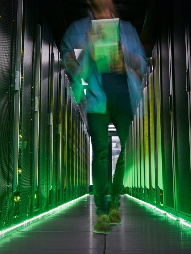 A person walks down a narrow aisle between tall server racks in a data center. The motion blur suggests fast movement or busy activity in the high-tech environment. Green lighting and indicator LEDs create a futuristic glow along the floor and equipment. The scene emphasizes modern infrastructure, digital networks, and information technology operations.