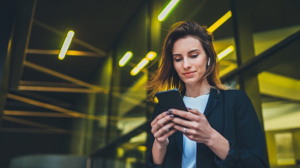 Lifestyle portrait of happy business woman walking in evening city and using mobile gadget, beautiful young girl banker dressed in business style listening to music wireless earphones and typing text message on smartphone device, flare light