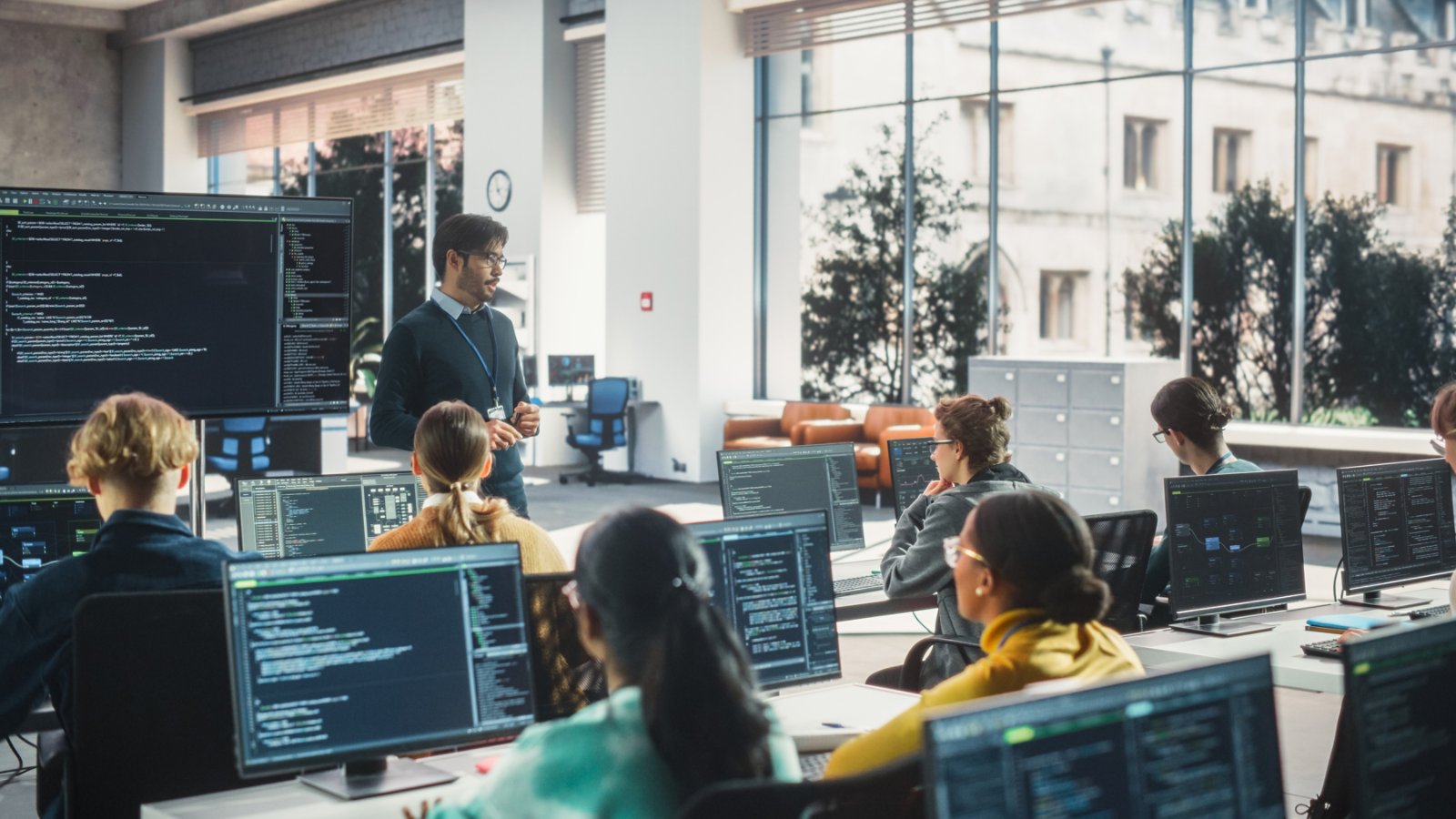A teacher stands at the front of a computer lab, addressing a group of students seated at desks with large monitors. The screens display lines of code and development environments, suggesting a programming or software engineering lesson. The classroom features large windows, bright natural light, and contemporary office furnishings. The scene conveys a collaborative, focused atmosphere in a modern educational or training setting.