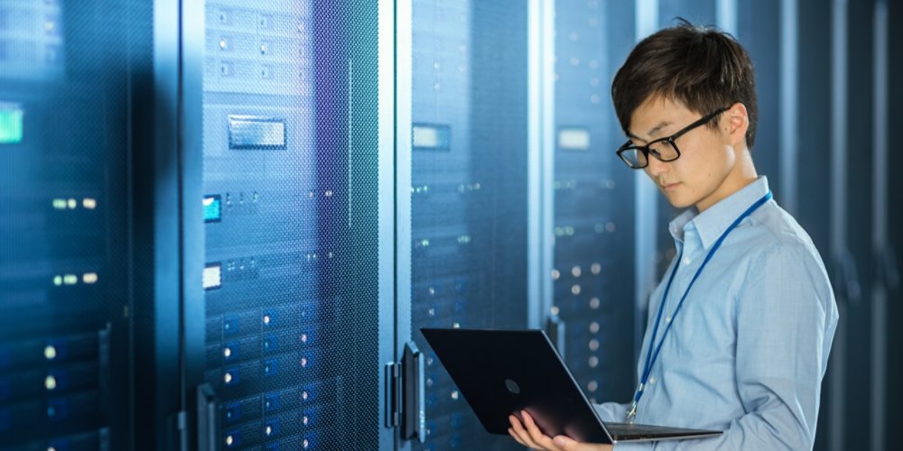 In the Modern Data Center: IT Engineer Standing Beside Server Rack Cabinets, Does Wireless Maintenance and Diagnostics Procedure with a Laptop.