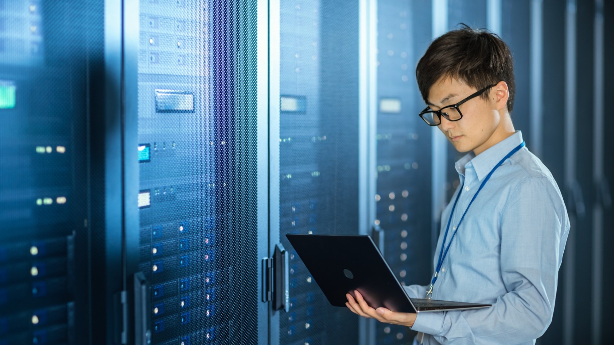 In the Modern Data Center: IT Engineer Standing Beside Server Rack Cabinets, Does Wireless Maintenance and Diagnostics Procedure with a Laptop.