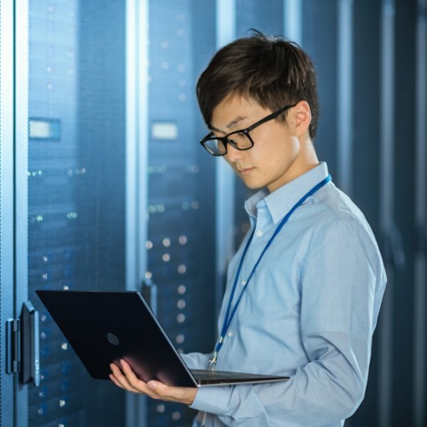In the Modern Data Center: IT Engineer Standing Beside Server Rack Cabinets, Does Wireless Maintenance and Diagnostics Procedure with a Laptop.