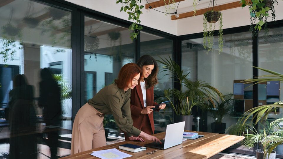 Smiling professional ladies entrepreneurs partners using laptop and tablet technology devices standing at work desk. Two happy business women of young and middle age talking working in green office.