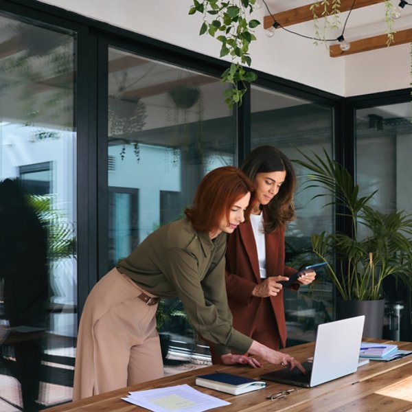 Smiling professional ladies entrepreneurs partners using laptop and tablet technology devices standing at work desk. Two happy business women of young and middle age talking working in green office.