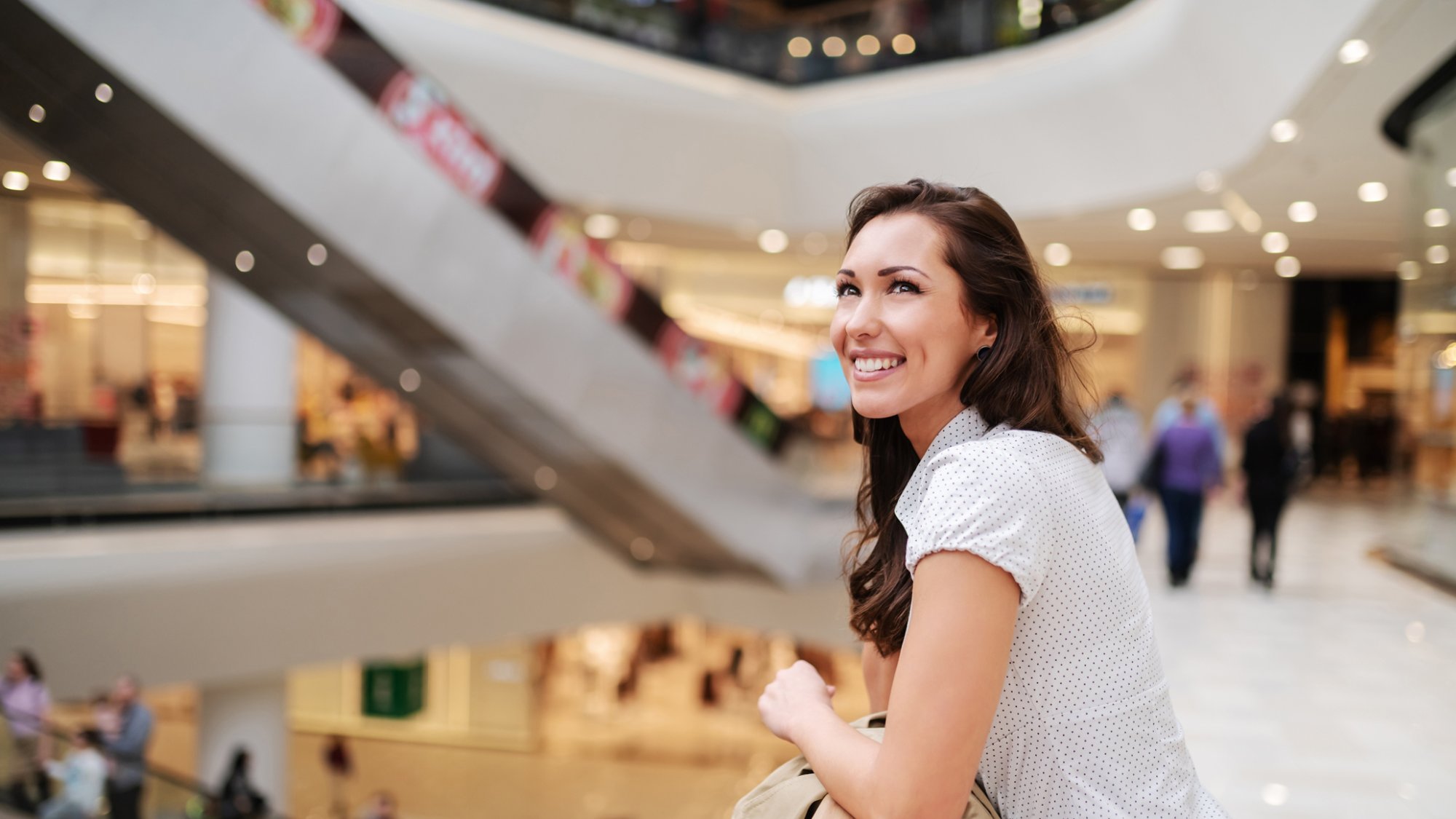 Beautiful caucasian brunette with stunning toothy smile leaning on railing and looking up. Shopping mall interior.