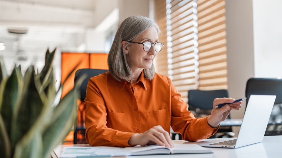 Portrait of beautiful confident senior woman, manager using laptop computer working online sitting in modern office. Business woman wearing stylish eyeglasses checking email. Technology concept 