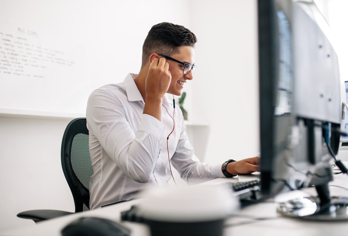 Smiling software developer sitting at his office desk working on laptop wearing earphones. Man wearing spectacles working on laptop computer in office.