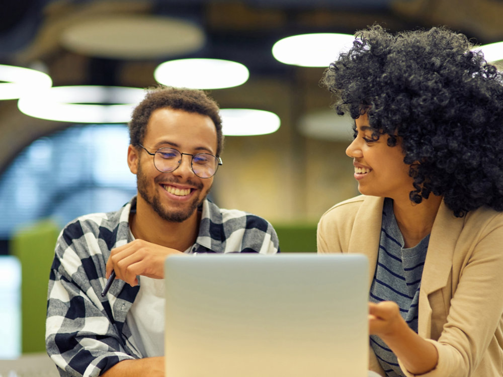 Two young happy multiracial business people sitting at the desk in the modern office, using laptop and discussing project, man and woman working together in coworking space. Office life and business