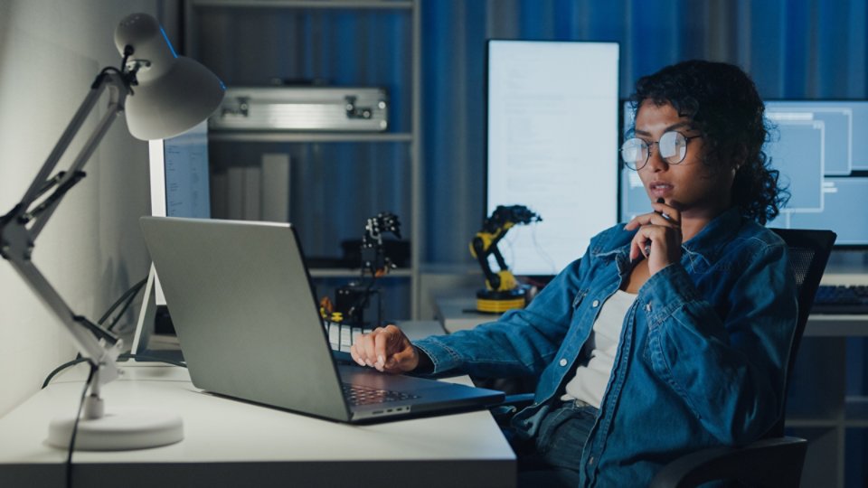 Young African American female programmer coding AI software on multiple screens in night office. Network security, cybersecurity, machine learning, working late, overtime, robotics automation concept.
