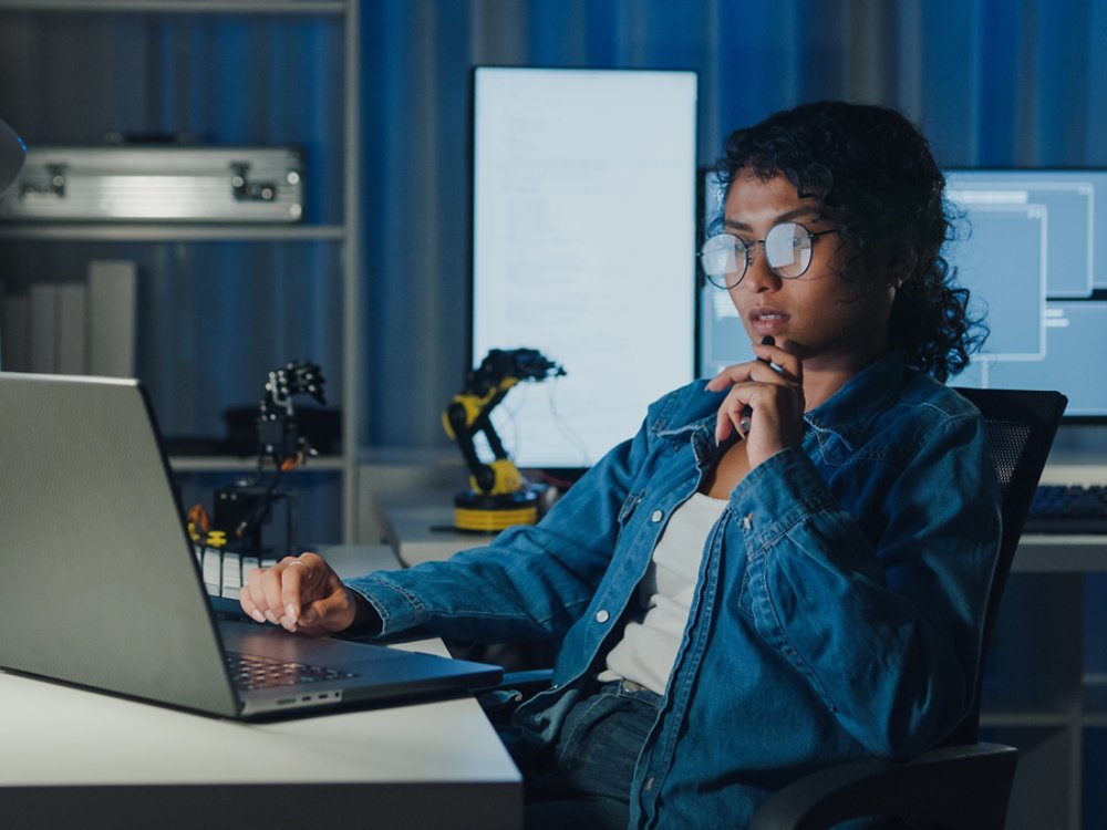 Young African American female programmer coding AI software on multiple screens in night office. Network security, cybersecurity, machine learning, working late, overtime, robotics automation concept.