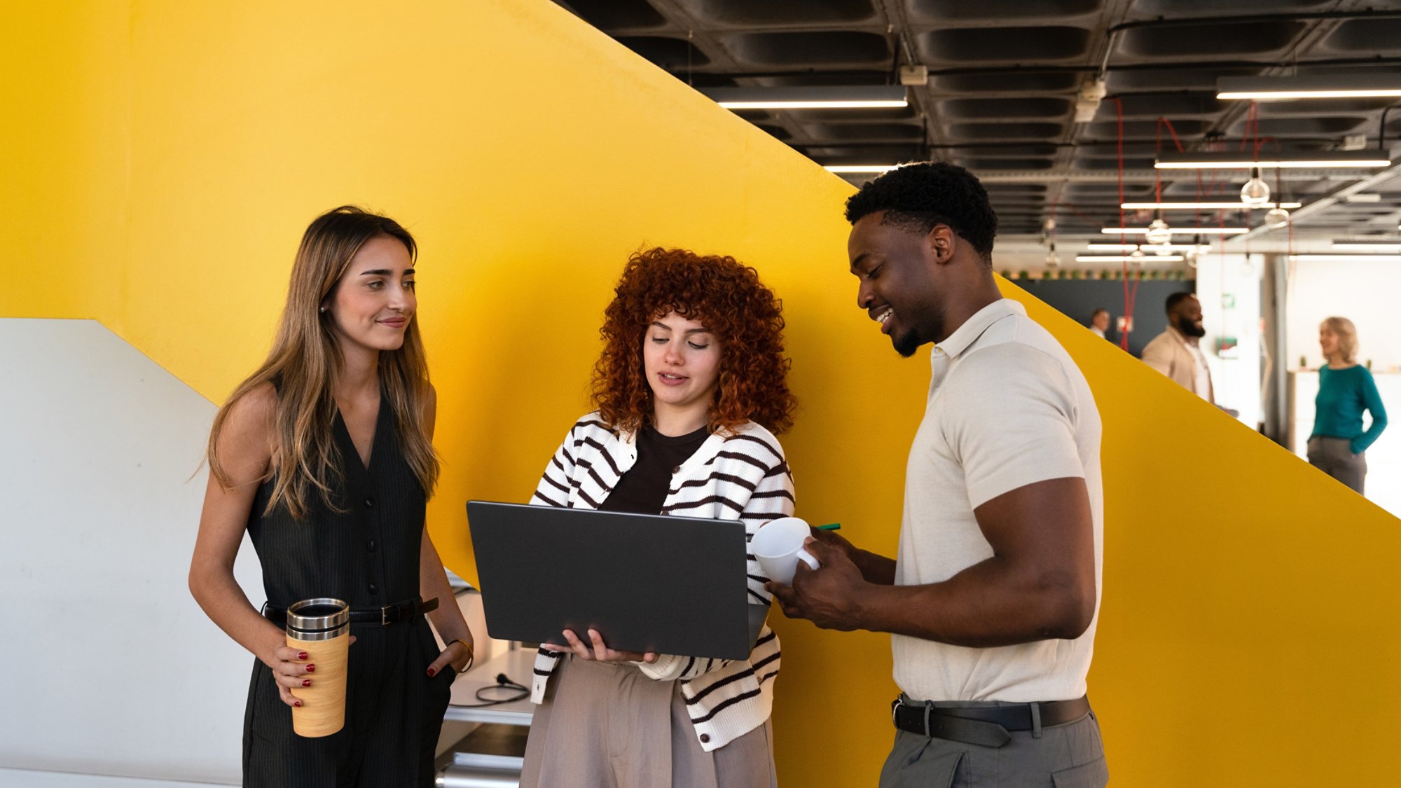 A group of diverse professionals engage in a lively conversation in a modern office, marked by a bright yellow wall. Collaborative teamwork and innovation are highlighted in this creative space.
