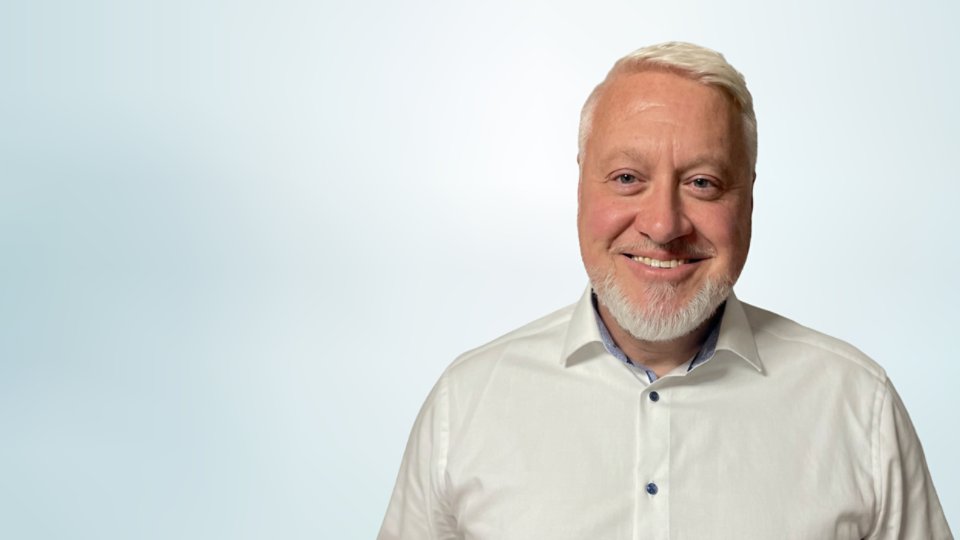 Portrait of a smiling adult man with short light hair and a trimmed beard, wearing a white button-up shirt. He is positioned on the right side of the frame against a soft light blue gradient background. The image has a clean, minimal look suitable for corporate, business, or professional profile use. Neutral lighting and simple composition emphasize the subject’s friendly expression.