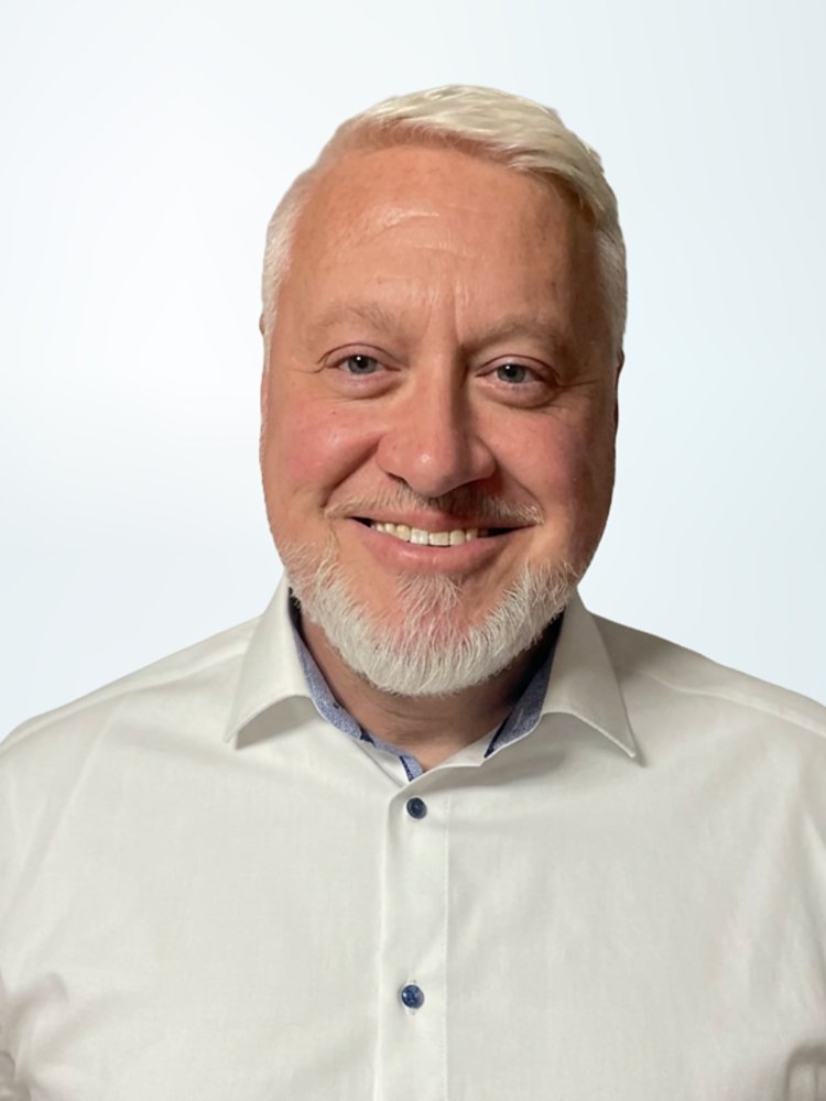 Portrait of a smiling adult man with short light hair and a trimmed beard, wearing a white button-up shirt. He is positioned on the right side of the frame against a soft light blue gradient background. The image has a clean, minimal look suitable for corporate, business, or professional profile use. Neutral lighting and simple composition emphasize the subject’s friendly expression.