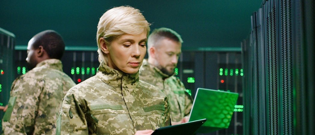 Caucasian military female officer in camouflage standing in server room with tablet device and checking data on monitors. Work with secret surveillance in army. Digital concept. Mixed-races men behind