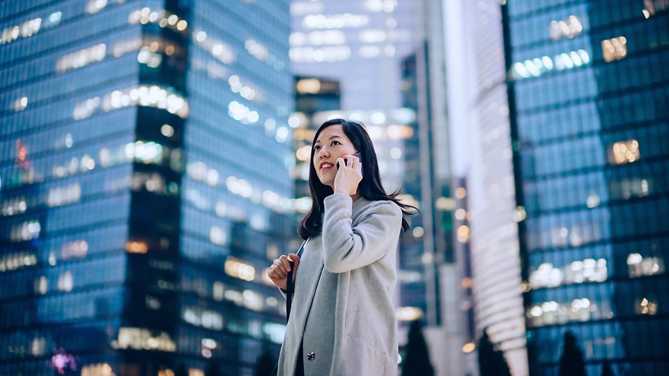 Confident and successful young Asian businesswoman talking on smartphone with business partners on the go, against contemporary corporate skyscrapers with illuminated facade in financial district in the evening. Business on the go