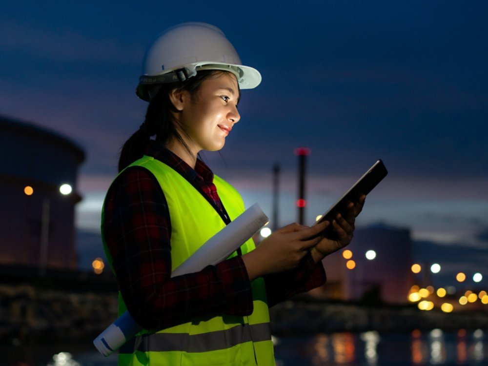 Asian woman petrochemical engineer working at night with digital tablet Inside oil and gas refinery plant industry factory at night for inspector safety quality control.
