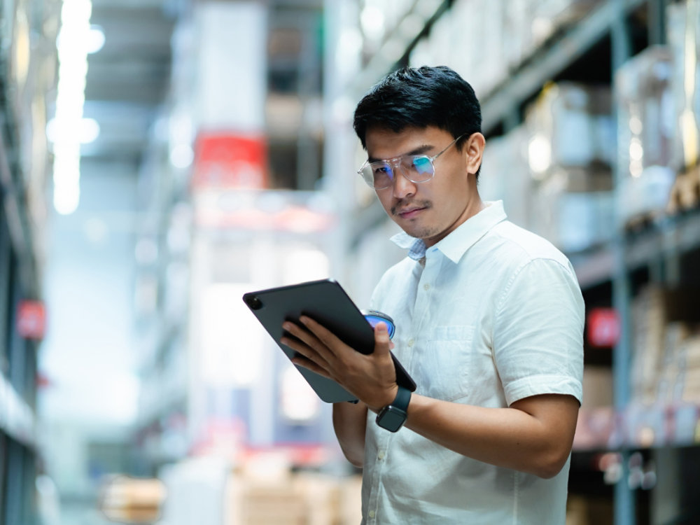 A man wearing glasses is looking at a tablet in a warehouse. He is focused on the screen and he is working