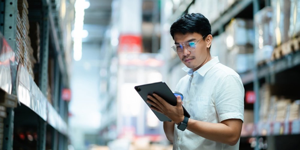 A man wearing glasses is looking at a tablet in a warehouse. He is focused on the screen and he is working