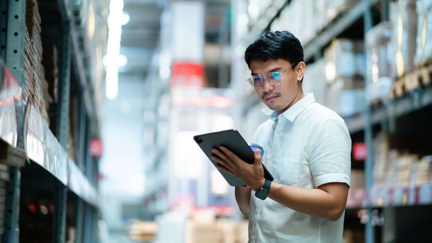 A man wearing glasses is looking at a tablet in a warehouse. He is focused on the screen and he is working