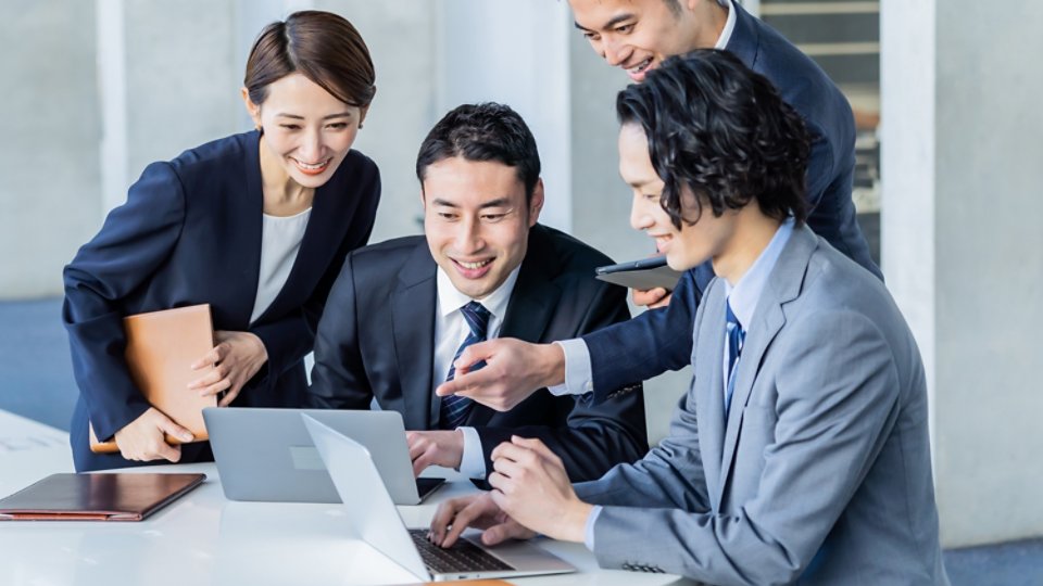 A group of professionals gathered around a table, engaging in a collaborative discussion. The setting appears to be a modern office environment with laptops and documents visible. The individuals are dressed in formal business attire, suggesting a corporate or professional meeting. The focus is on teamwork and communication.