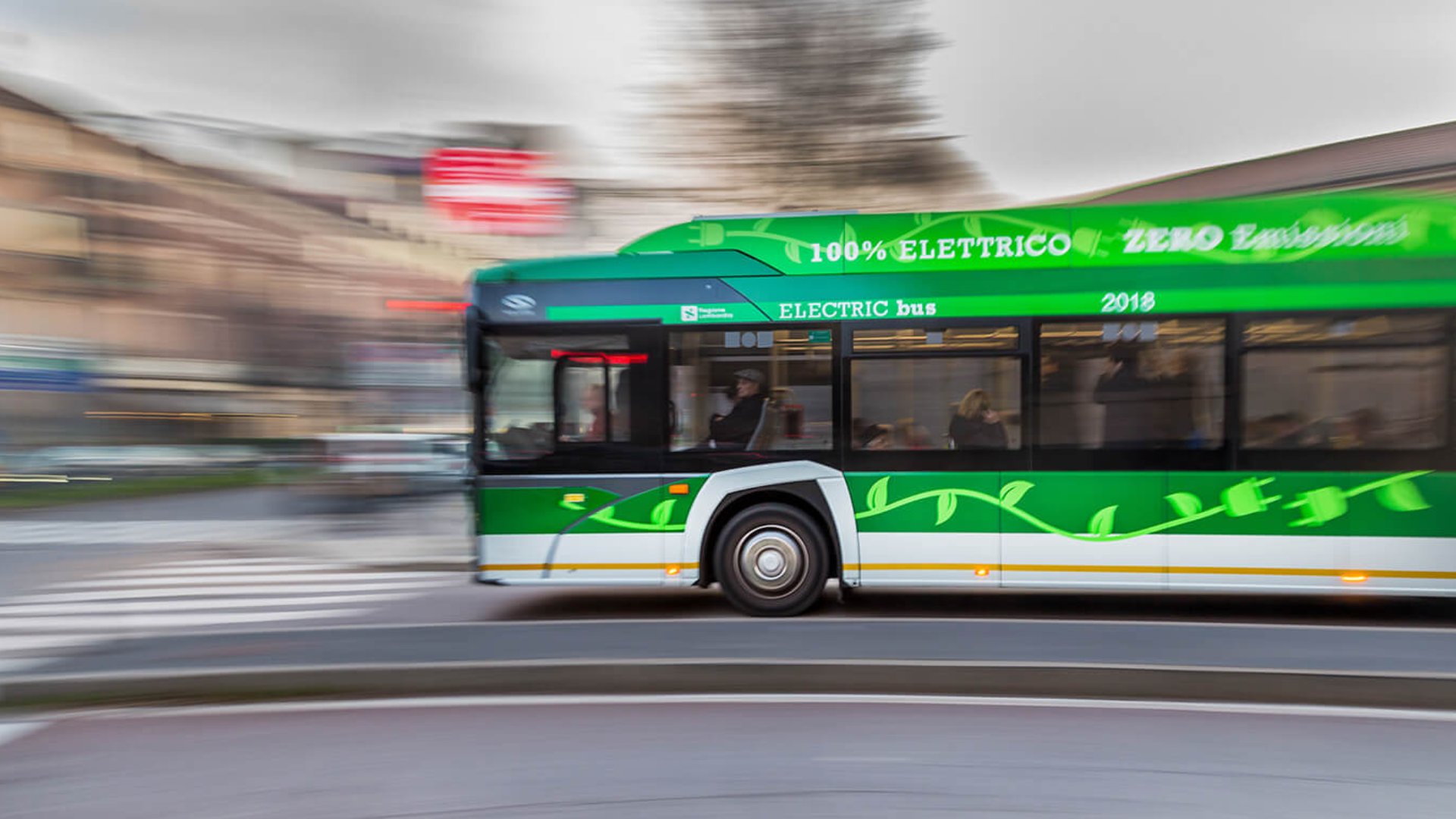 A green electric bus speeds along an urban street, captured with motion blur to emphasize movement. The side of the bus displays text including "100% ELETTRICO" and "ZERO Emission" along with "ELECTRIC bus" and the year "2018". Leaf graphics decorate the lower part of the vehicle, reinforcing its eco-friendly theme. Buildings and a crosswalk in the background appear blurred, highlighting the bus as the main subject.