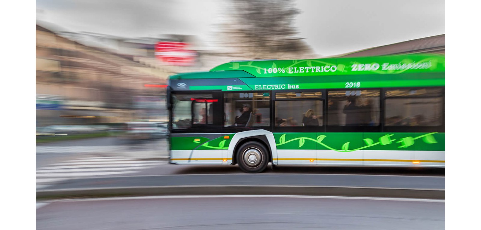 A green electric bus speeds along an urban street, captured with motion blur to emphasize movement. The side of the bus displays text including "100% ELETTRICO" and "ZERO Emission" along with "ELECTRIC bus" and the year "2018". Leaf graphics decorate the lower part of the vehicle, reinforcing its eco-friendly theme. Buildings and a crosswalk in the background appear blurred, highlighting the bus as the main subject.