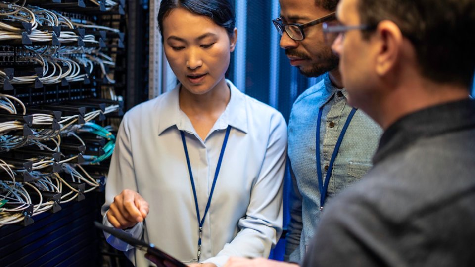 Three IT technician looking at a digital tablet and talking while standing next to a server in a data center.
