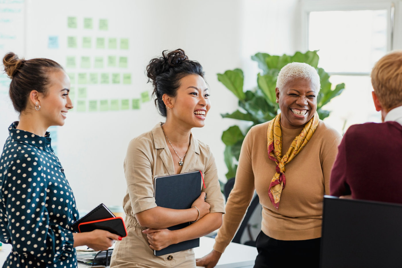 Colleagues standing in a small group discussing something while laughing. Two of the women are holding notebooks.