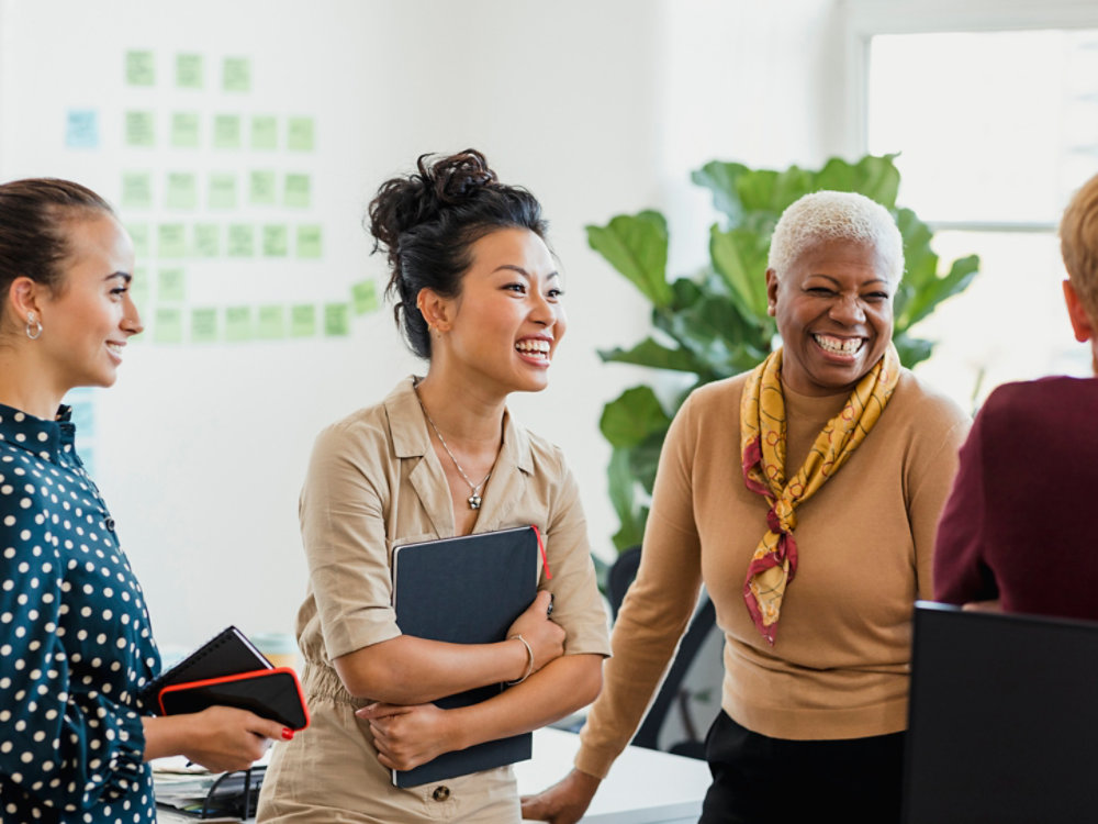 Colleagues standing in a small group discussing something while laughing. Two of the women are holding notebooks.