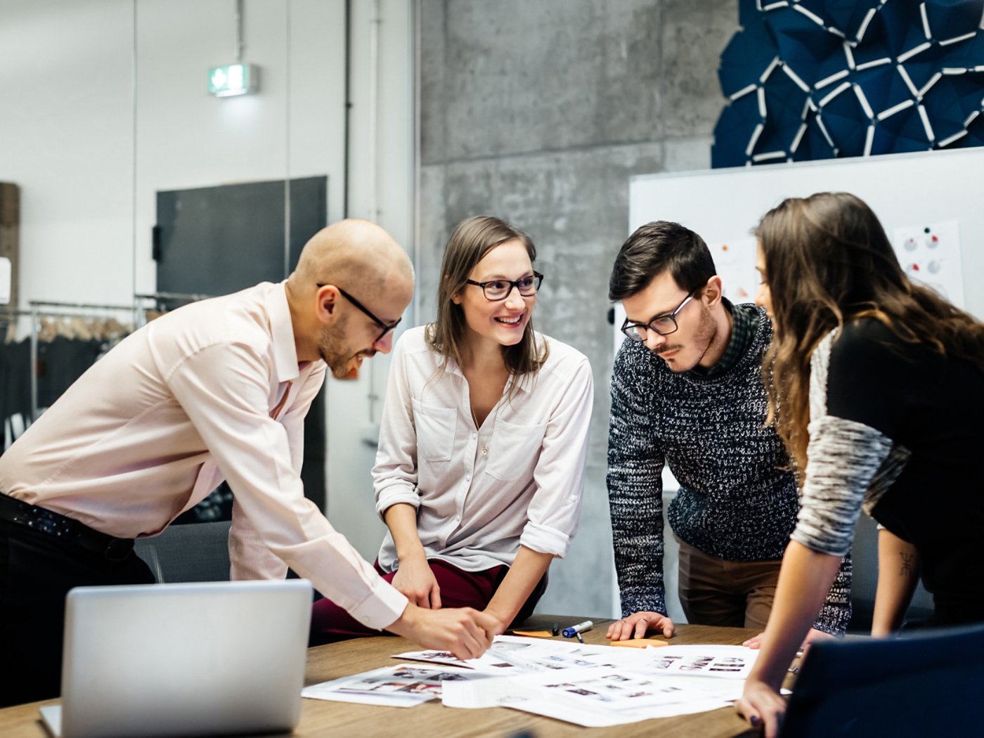 A team of four business people is standing in front a desk in a bright office room. They ambitiously point at documents while smiling and looking at the charts and notes.