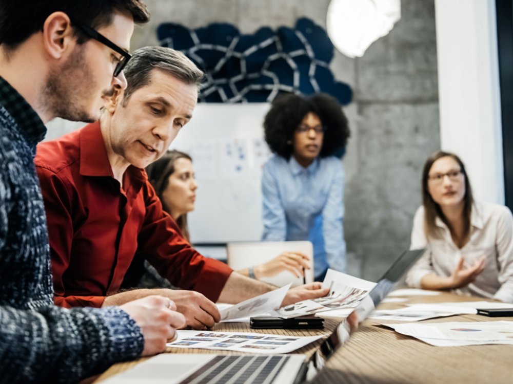 Two men are looking at documents in a business meeting. Women can be seen in the background talking to someone else.