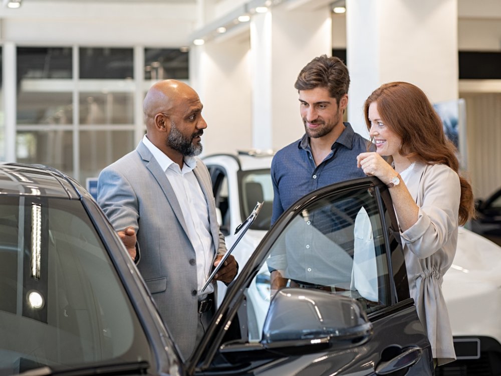 Beautiful couple in casual clothes smiling while examining a new car in a motor show with salesman. Mature salesman showing new luxury car to man and woman in showroom. Happy couple with cardealer showing new features in auto dealership.