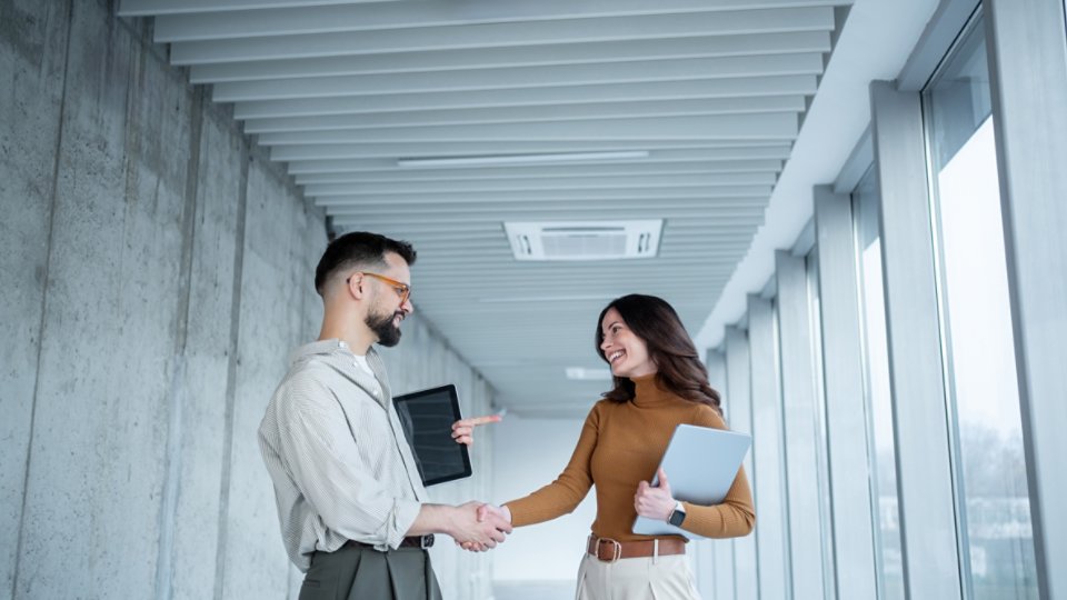 Two young professionals stand in a bright office corridor, shaking hands and smiling at each other. The man holds a tablet while the woman carries a laptop and points toward him in a friendly, conversational gesture. Large windows line the hallway, letting in natural light and creating a clean, contemporary business environment. The overall mood is positive and collaborative, suggesting a successful meeting or new partnership.