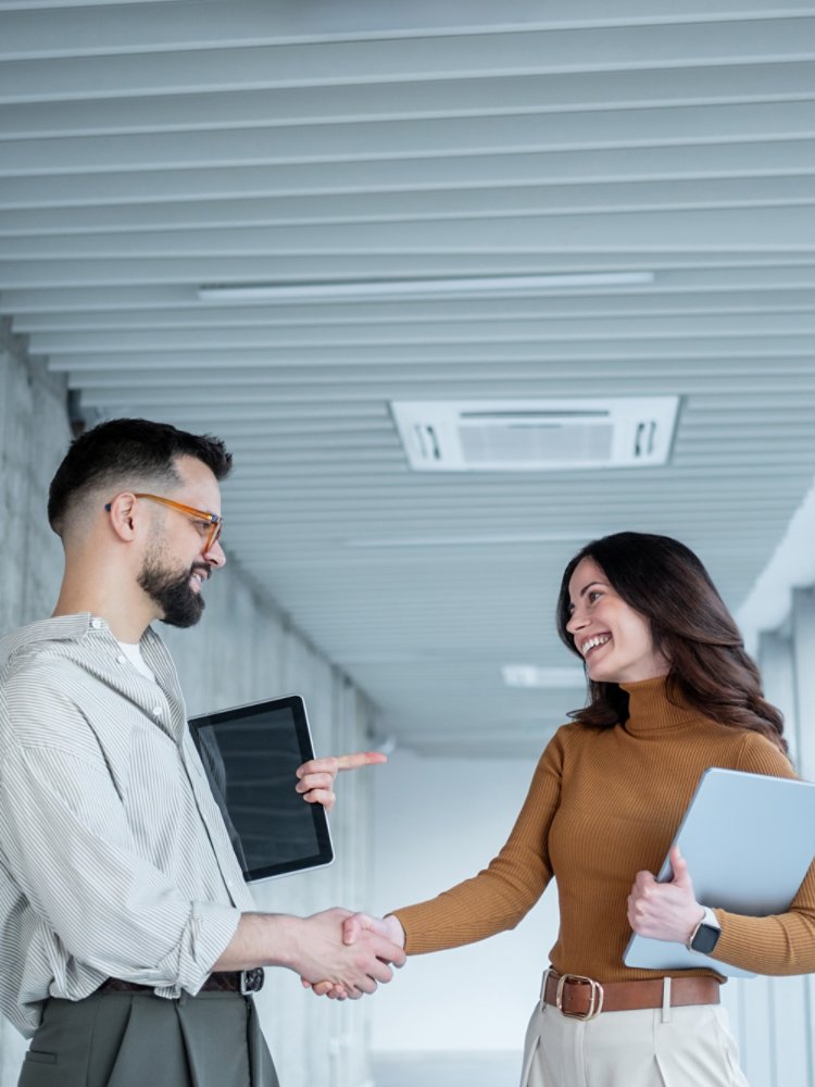Two young professionals stand in a bright office corridor, shaking hands and smiling at each other. The man holds a tablet while the woman carries a laptop and points toward him in a friendly, conversational gesture. Large windows line the hallway, letting in natural light and creating a clean, contemporary business environment. The overall mood is positive and collaborative, suggesting a successful meeting or new partnership.