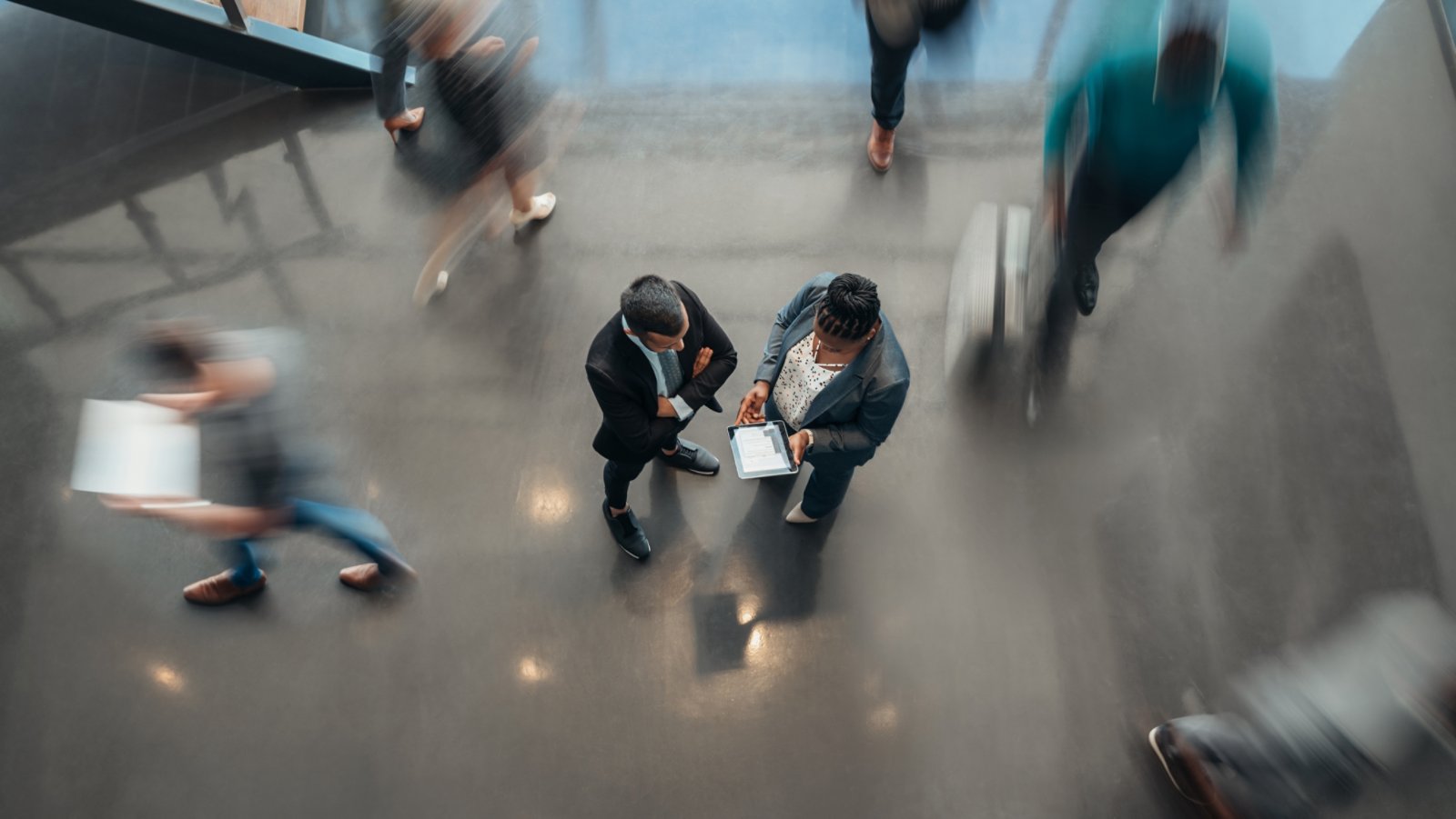 Two business people standing in the lobby of an office looking at a tablet while people are walking past in a blur