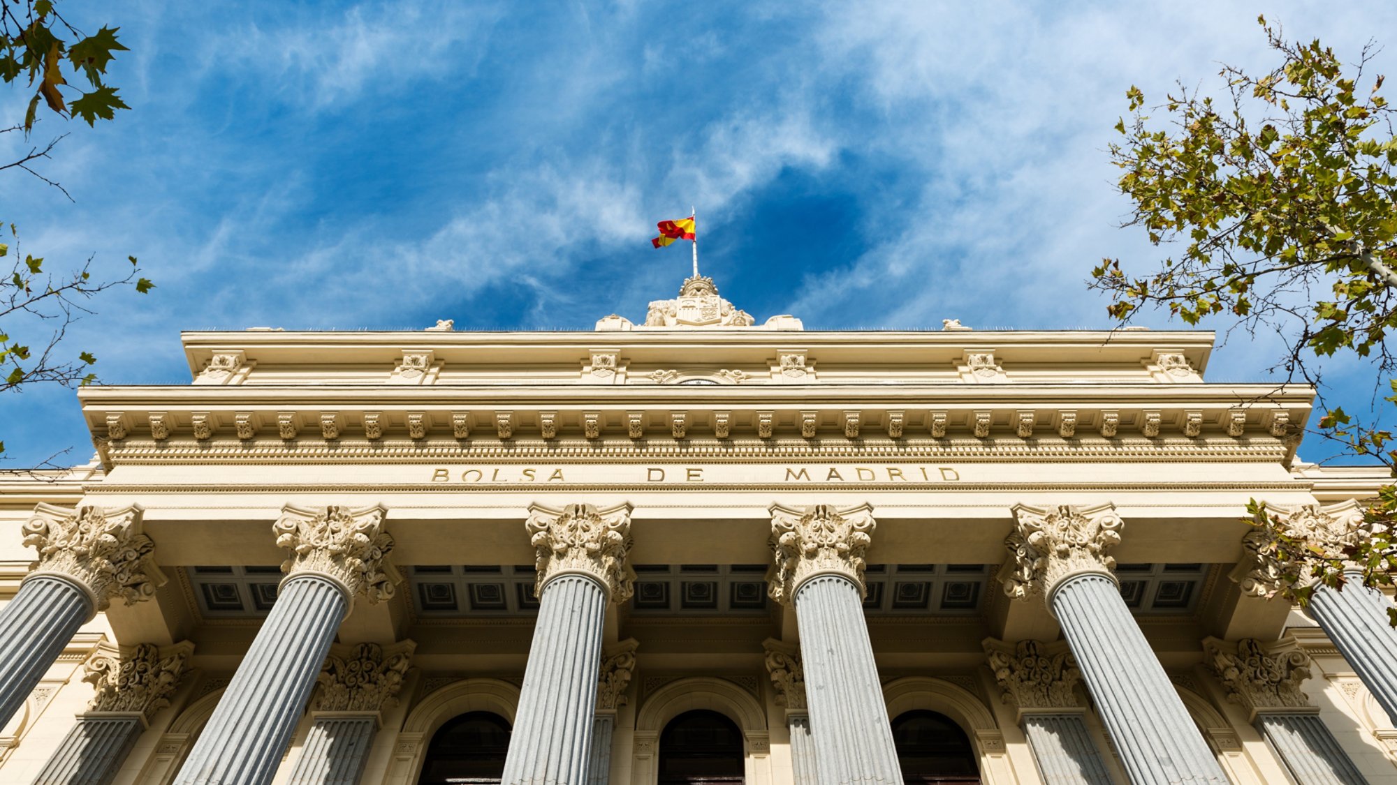 Low-angle view of the columns and neoclassical facade of the Bolsa de Madrid (Madrid's Stock Exchange) against a cloudy blue sky.