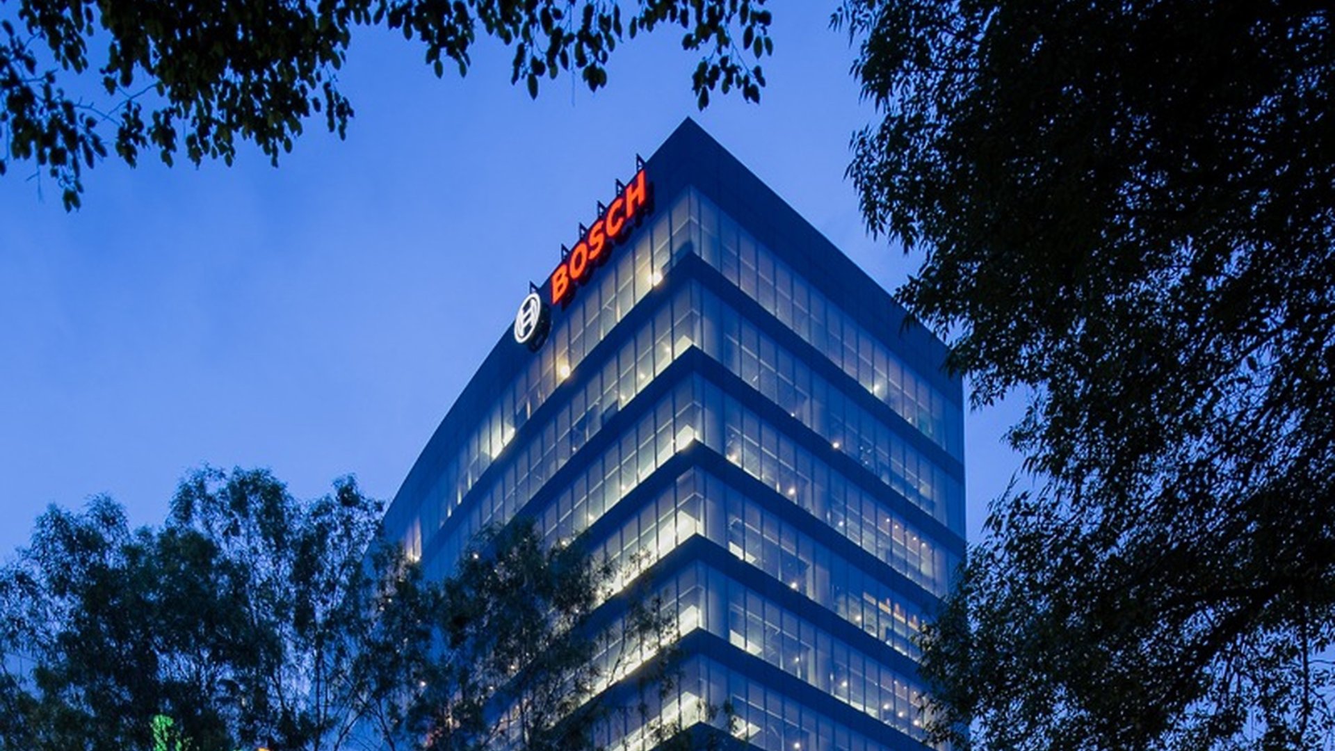 A modern glass office tower rises above surrounding trees in the early evening. The building is illuminated from within, with rows of lights visible across multiple floors. The Bosch name and circular emblem are clearly visible in bright red and white near the top of the structure. The sky is a deep blue, creating a calm urban atmosphere framed by dark foliage.