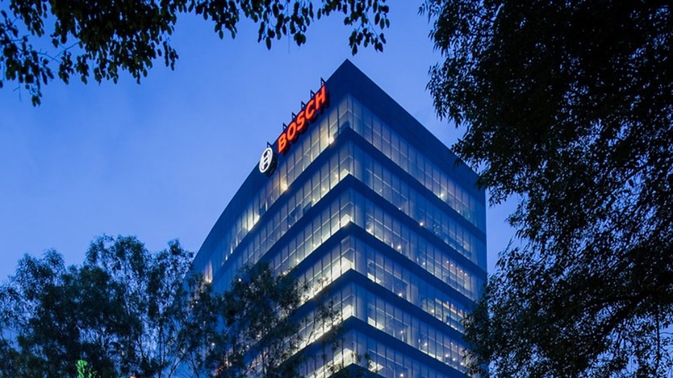 A modern glass office tower rises above surrounding trees in the early evening. The building is illuminated from within, with rows of lights visible across multiple floors. The Bosch name and circular emblem are clearly visible in bright red and white near the top of the structure. The sky is a deep blue, creating a calm urban atmosphere framed by dark foliage.