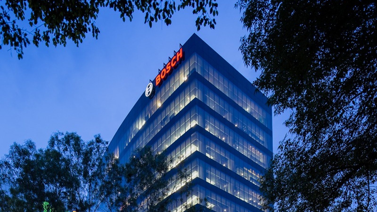 A modern glass office tower rises above surrounding trees in the early evening. The building is illuminated from within, with rows of lights visible across multiple floors. The Bosch name and circular emblem are clearly visible in bright red and white near the top of the structure. The sky is a deep blue, creating a calm urban atmosphere framed by dark foliage.
