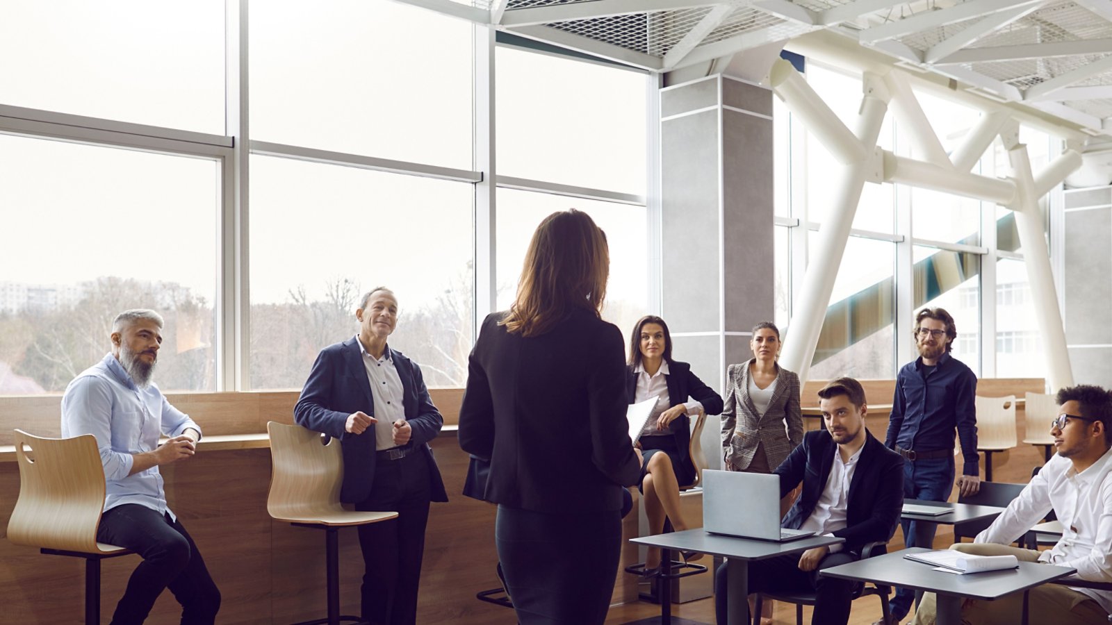 Various businessmen listen to female business leader during corporate meeting or training lecture. Rear view of woman in business attire speaking to people in modern business center. Panoramic view.