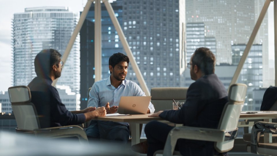 Three professionals sit around a conference table in a high-rise office, engaged in a business discussion. The central figure uses a laptop while the others listen and converse, suggesting a collaborative meeting or presentation. Large windows reveal a contemporary city skyline with tall buildings and glass architecture, adding an urban corporate atmosphere. The lighting is soft and natural, creating a focused yet relaxed professional mood.